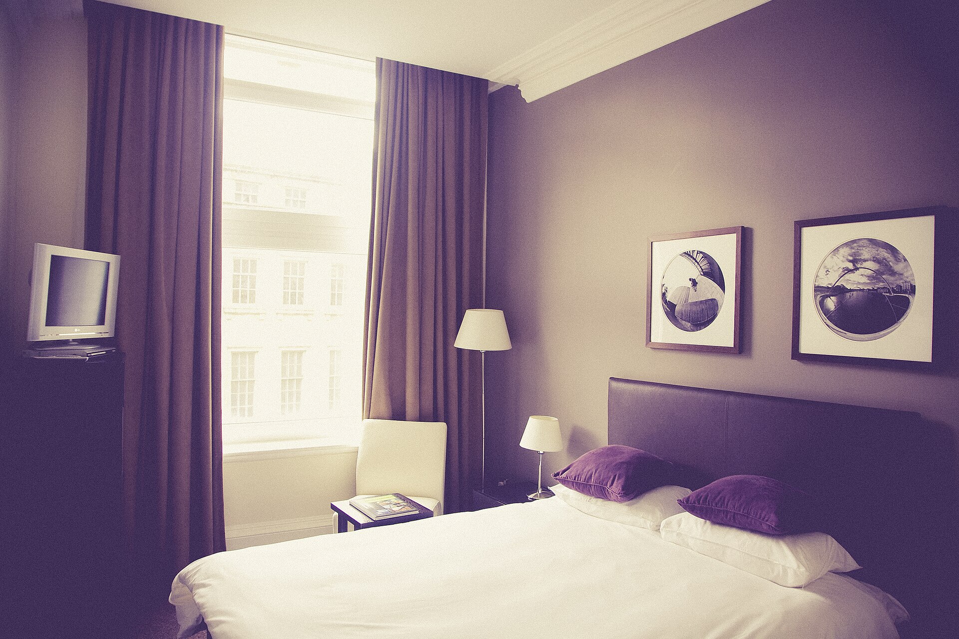 Hotel-style bedroom with a made double bed, framed prints and tall windows with purple curtains