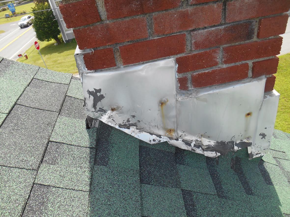 Close-up of worn, poorly sealed metal flashing where a brick chimney meets asphalt roof shingles