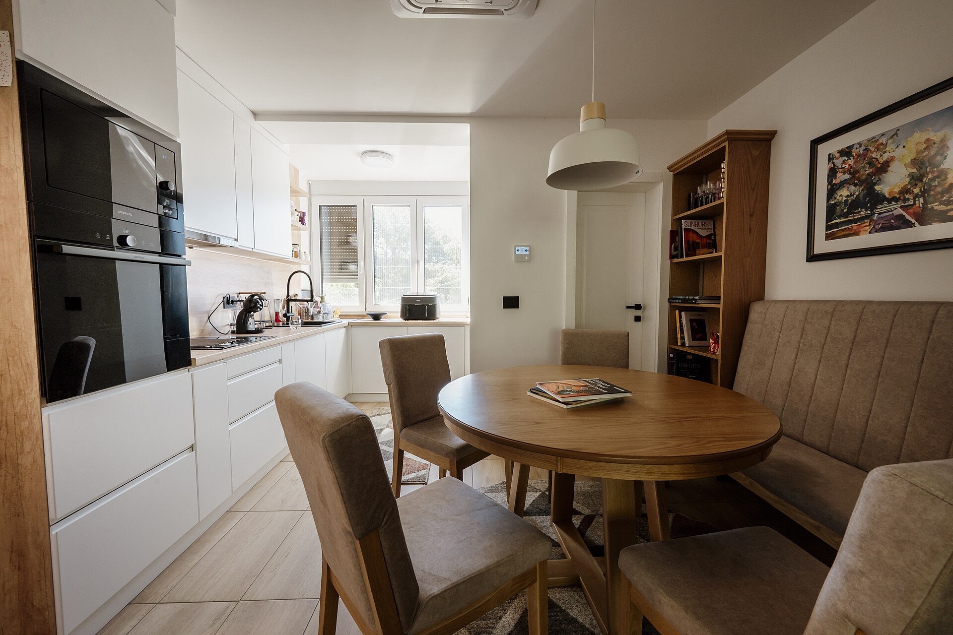 Bright white kitchen with built-in oven and light wood floor opening onto a round wooden dining table
