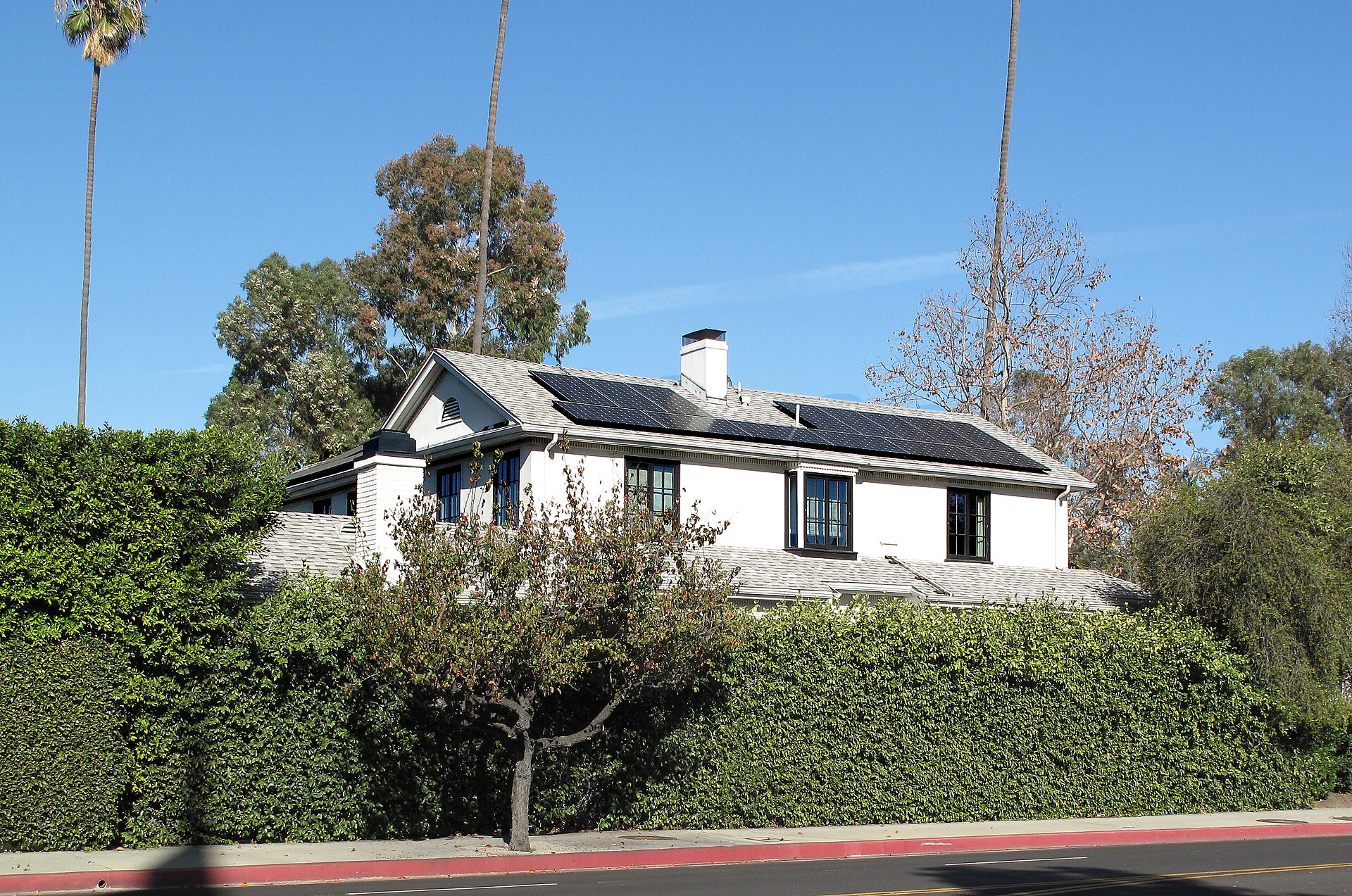 White one-storey house with dark trim and a row of solar panels on its low-pitched roof behind a hedge